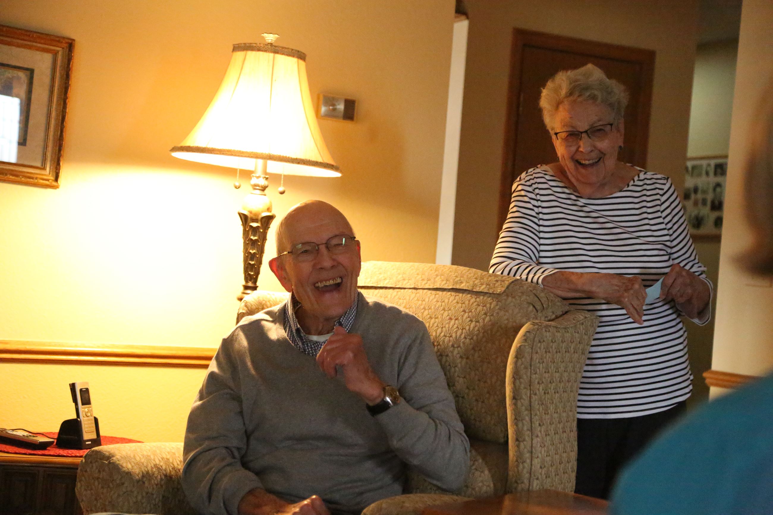 Jim Sparks sits laughing in chair with wife standing behind him smiling